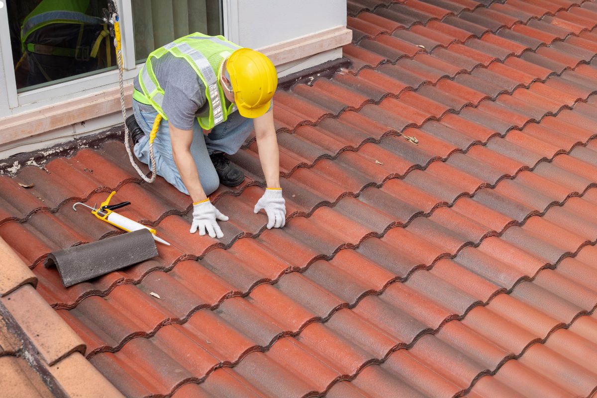 A roofer in a hard hat and yellow vest performs a roof inspection, checking for safety and structural integrity.