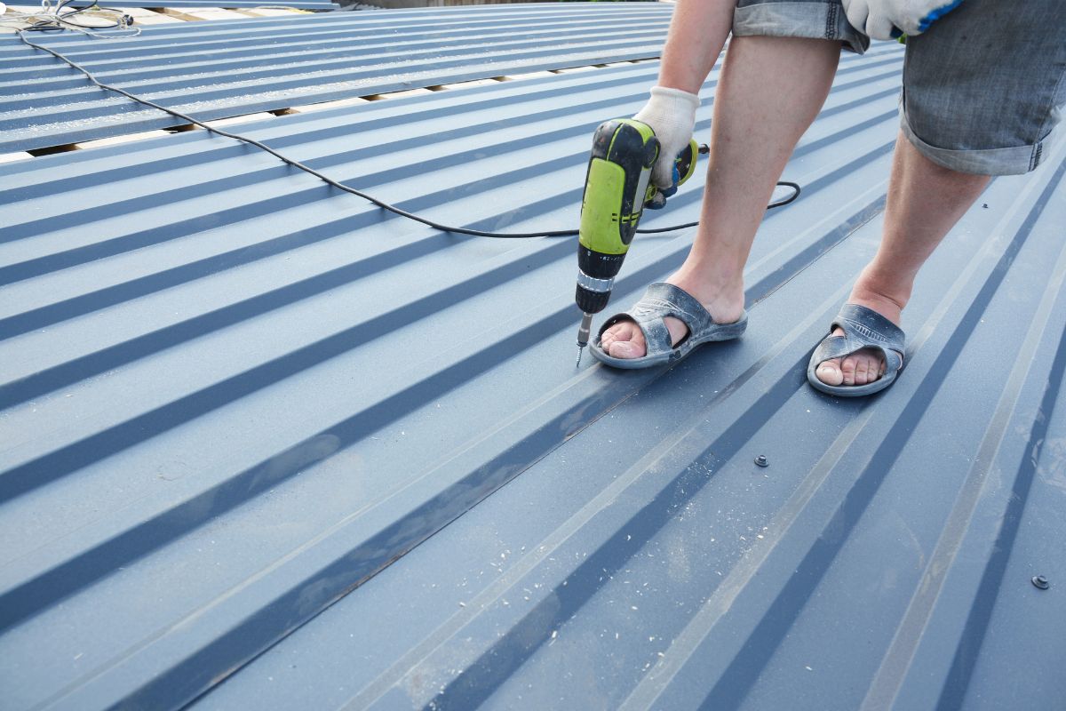 A person with sandals is on a roof, actively using a drill for maintenance or construction purposes.