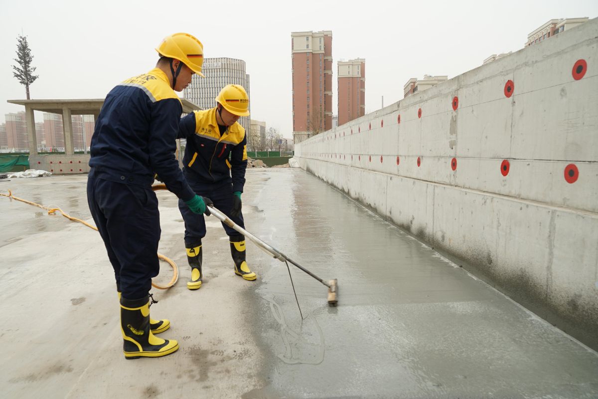 Two workers in yellow helmets and boots apply wet concrete to a smooth surface at a construction site. Buildings are visible in the background.