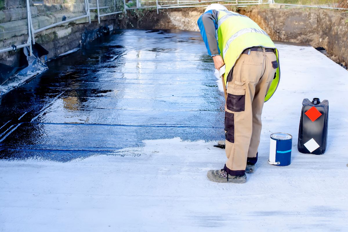 Two workers in yellow helmets and boots apply wet concrete to a smooth surface at a construction site. Buildings are visible in the background.
