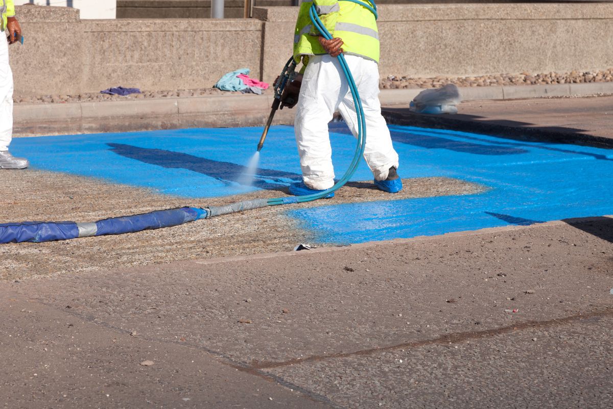 A worker in a bright yellow vest sprays blue paint onto a street surface, creating a vibrant blue area on the pavement.