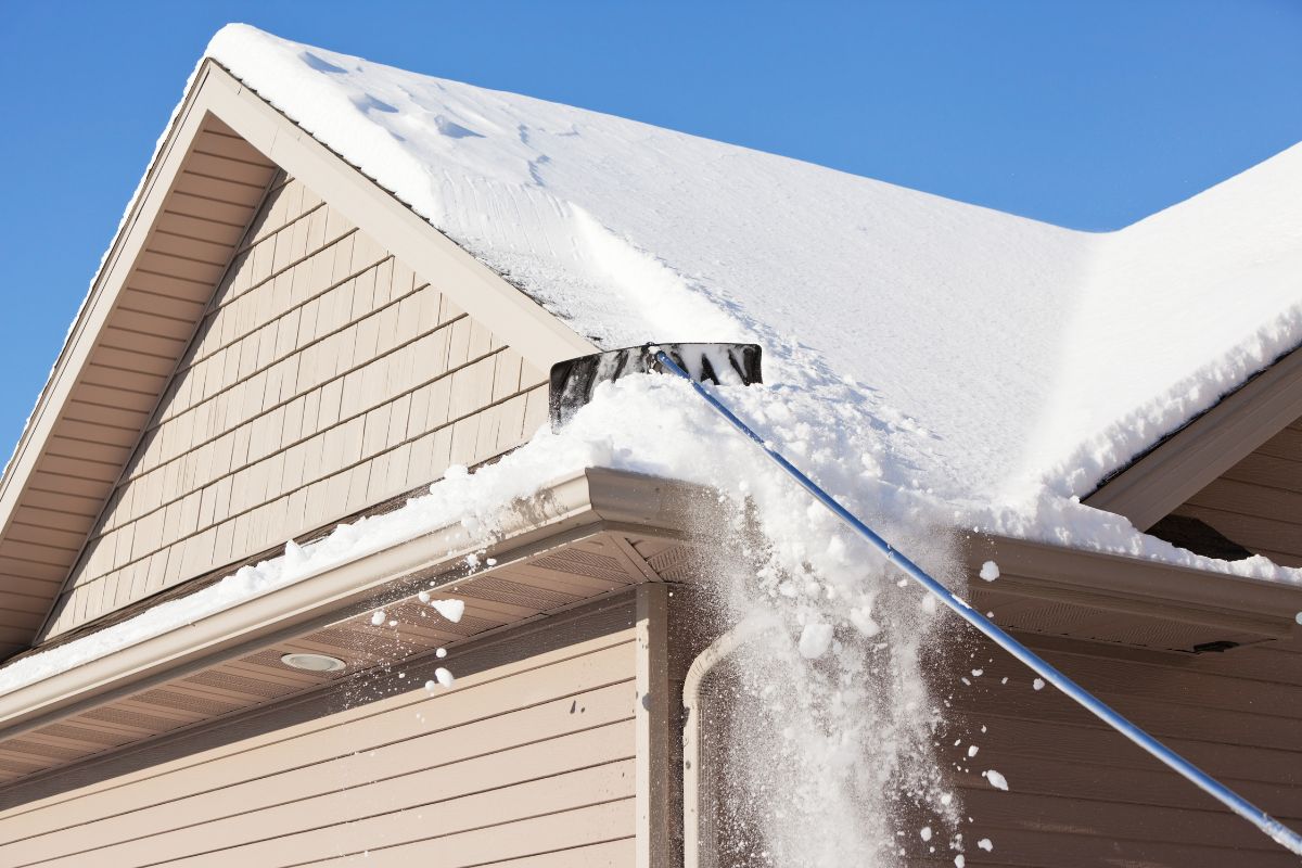 A person uses a long-handled snow shovel to clear snow off the roof of a house on a sunny winter day.