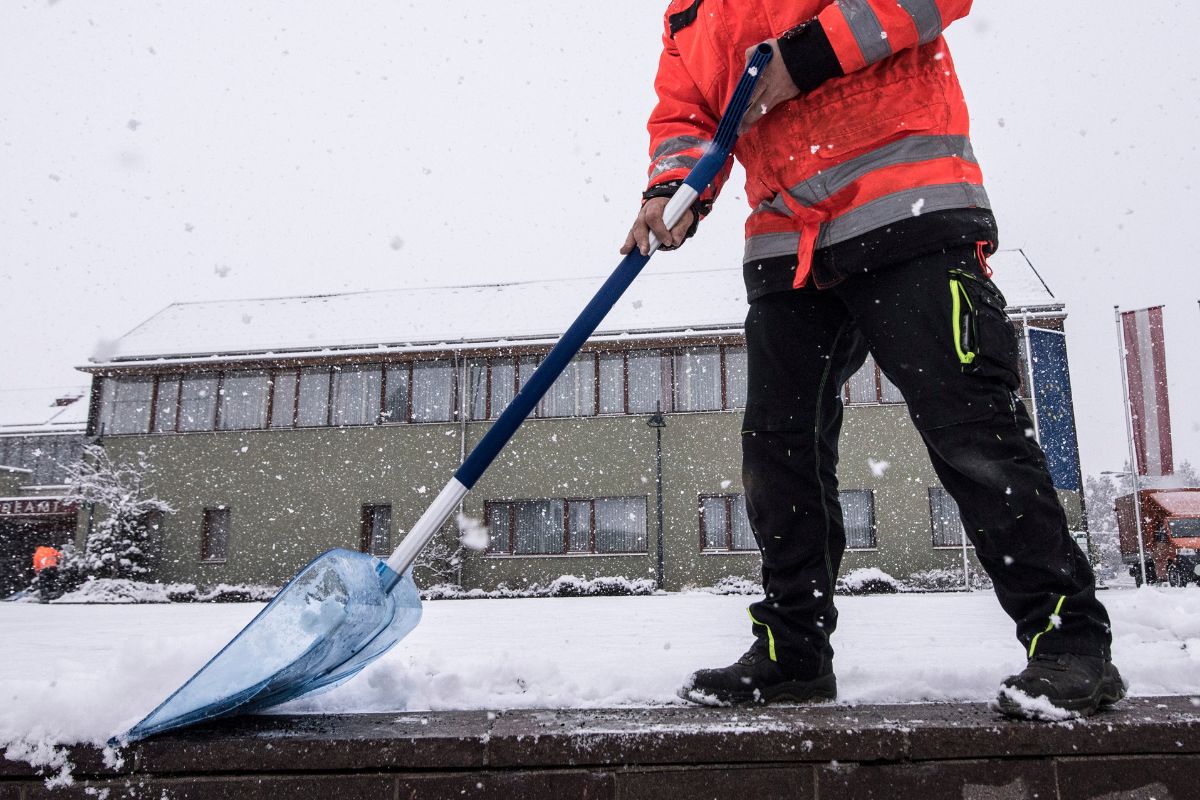 A worker in an orange jacket shovels snow from a sidewalk, with a snowy building and heavy snowfall in the background.