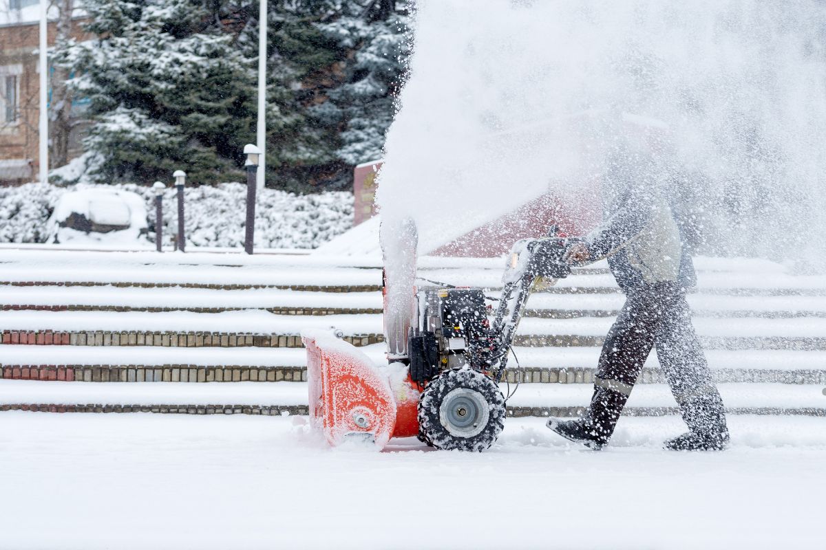 A person operates a snowblower, clearing a snowy path while snowflakes swirl around them in a winter landscape.